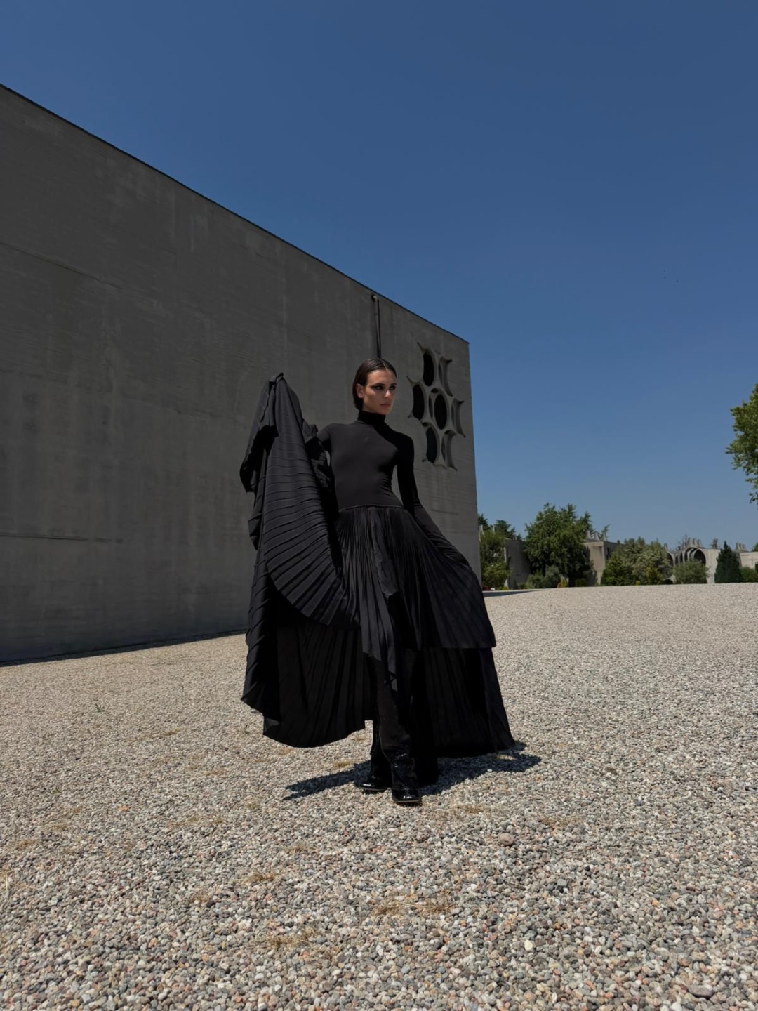 Pleated black dress and jacket against brutalist architecture, gravel.