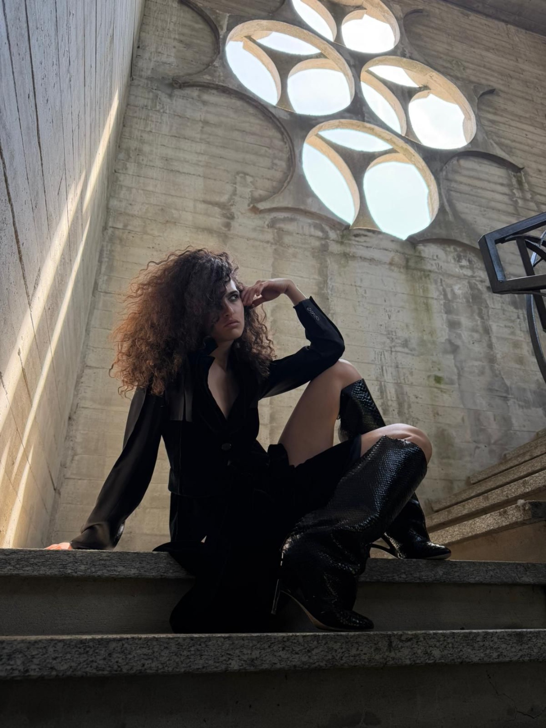 A figure with curly hair in velvet and sheer, seated under a chapel rosette window.