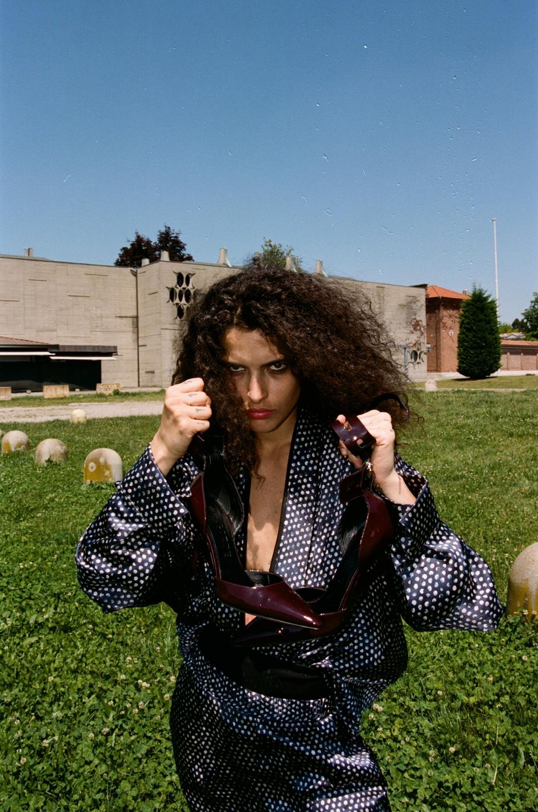 Polka-dot silk suit, fists raised, holding shoes in a grass field.